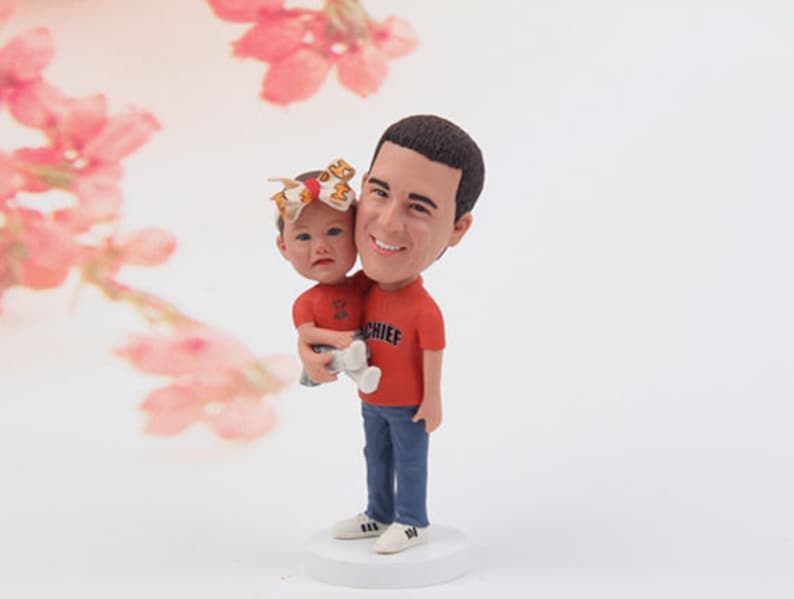 May include: A personalised figurine of a man holding a small girl. The man is wearing a red shirt with the word "Chief" on it and blue jeans. The girl is wearing a white shirt and blue jeans. The figurine is on a white base.