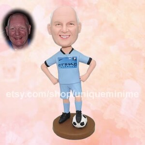 May include: A custom bobblehead figurine of a person wearing a light blue football shirt with the text "ETIHAD AIRWAYS" on the front. The figurine is holding a football and standing on a brown base.