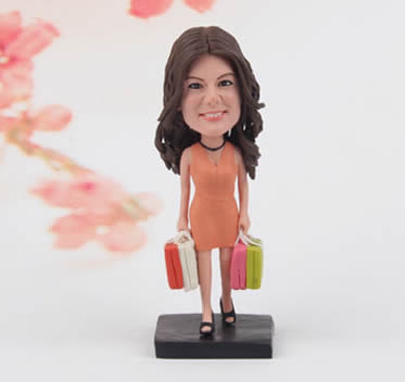 May include: A custom caricature figurine of a woman with brown hair, wearing a peach dress and holding two shopping bags. The figurine is standing on a black base.