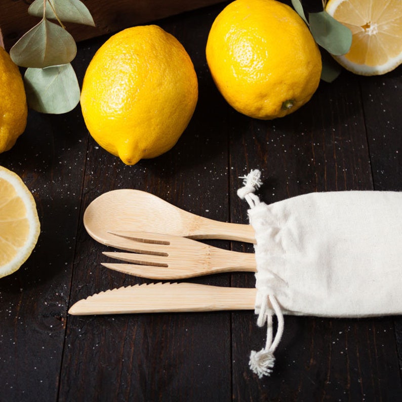 May include: A set of light-colored bamboo cutlery consisting of a spoon, a fork, and a serrated knife, partially tucked into a small, off-white fabric drawstring pouch. The utensils are arranged horizontally alongside several bright yellow lemons.