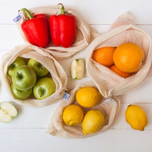 May include: Four reusable mesh produce bags filled with fresh produce. The bags are made of natural cotton and have a drawstring closure. The bags are filled with red bell peppers, green apples, oranges, and lemons. The bags are sitting on a white wooden surface.