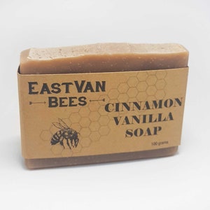 May include: A brown bar of soap with a label that reads "East Van Bees Cinnamon Vanilla Soap 100 grams". The label has a honeycomb pattern and a bee illustration.