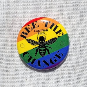 May include: A round button with a rainbow design and the text "BEE THE CHANGE" in black. A black silhouette of a bee is in the center of the button with the text "EASTVAN BEES" above it.