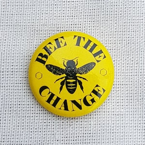 May include: Yellow button with a black bee and the text "Bee the Change" in black.