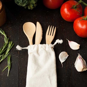 May include: A set of light-colored wooden cutlery consisting of a spoon, a serrated knife, and a fork, all housed within a plain, off-white fabric drawstring pouch. The utensils are arranged vertically with their handles tucked inside the bag.