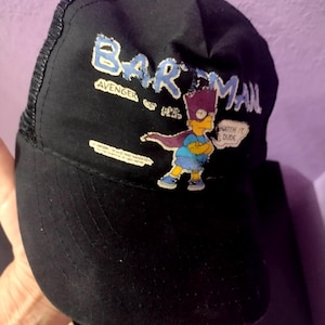 May include: Black baseball cap with a mesh back and a curved brim. The cap features a graphic of Bart Simpson as "Bartman" with the text "Bartman Avenger of the 90s" and "Watch it Dude".