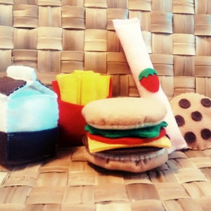 May include: A felt food set featuring a hamburger, french fries in a red container, a carton of milk, a cookie, and a ketchup packet with a strawberry. The items are made of felt in various colors.