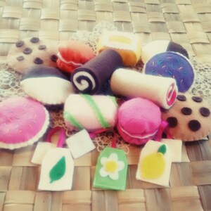 May include: A collection of colorful felt desserts, including cookies, macarons, and a swiss roll. The treats are arranged on a woven surface, with small felt tote bags featuring a leaf, flower, and lemon design.
