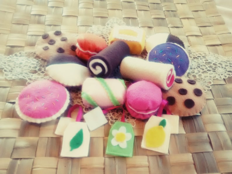 May include: A collection of colorful felt desserts, including cookies, macarons, and a swiss roll. The treats are arranged on a woven surface, with small felt tote bags featuring a leaf, flower, and lemon design.