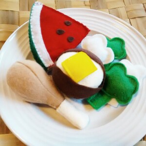May include: A felt food plate featuring a watermelon slice, a fried egg with a yellow square of butter, broccoli florets, and a chicken leg. The items are arranged on a white plate.
