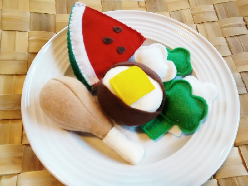 May include: A felt food plate featuring a watermelon slice, a fried egg with a yellow square of butter, broccoli florets, and a chicken leg. The items are arranged on a white plate.
