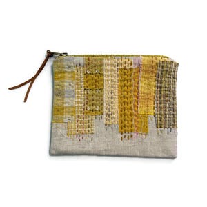 May include: A rectangular, handmade pouch with a patchwork design. The top section features a variety of yellow and olive-green woven fabrics, while the bottom is a neutral grey. A brown leather pull is attached to the zip.
