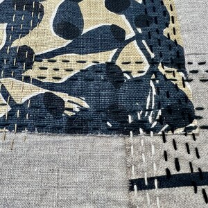 May include: A beige fabric with black stitching and a black and beige patterned fabric patch sewn on top. The patterned fabric has a design of abstract shapes.