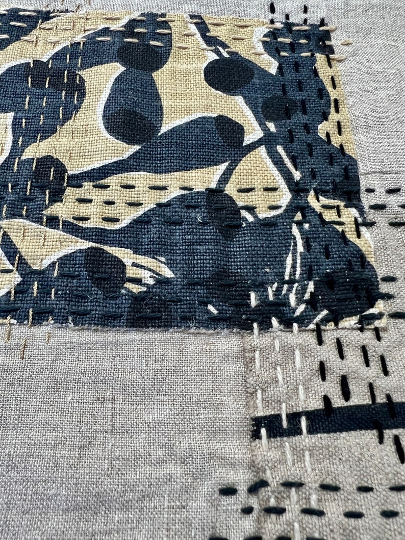 May include: A beige fabric with black stitching and a black and beige patterned fabric patch sewn on top. The patterned fabric has a design of abstract shapes.