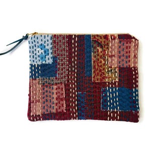 May include: A rectangular zippered pouch featuring a patchwork design of various fabric textures in shades of deep red, navy blue, light blue, and brown. The surface is covered in rows of white running stitches and includes a dark leather zipper pull.