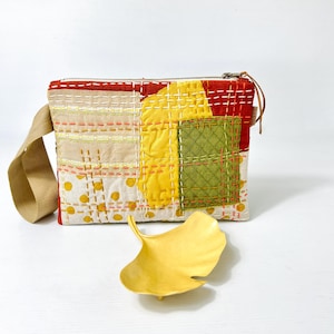 Könnte beinhalten: Eine Patchwork-Tasche mit Reißverschluss und einem braunen Trageriemen. Die Tasche besteht aus verschiedenen Stoffen in Rot-, Gelb-, Grün- und Weißtönen. Die Tasche ist mit Nähten und kleinen Punkten verziert. Im Vordergrund liegt ein gelbes Ginkgoblatt.