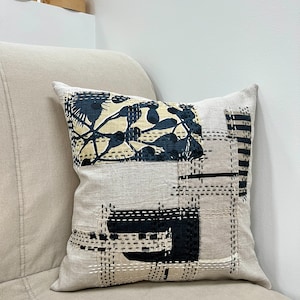 May include: A beige linen pillow with a black and white geometric design. The design features a large black floral pattern in the center and smaller black geometric shapes around the edges.