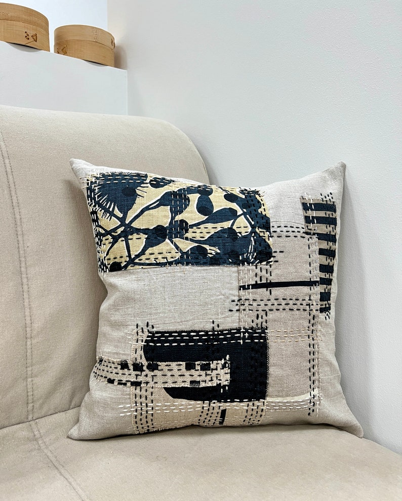May include: A beige linen pillow with a black and white geometric design. The design features a large black floral pattern in the center and smaller black geometric shapes around the edges.