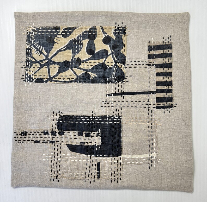 May include: A beige linen cushion with a black and white abstract design created with stitched fabric scraps. The design features geometric shapes and a floral pattern.