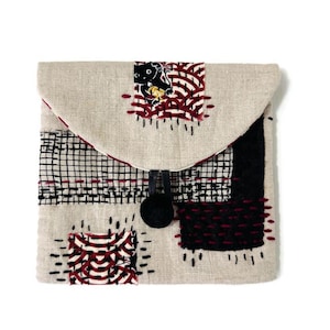 May include: A beige fabric pouch with a flap closure. The pouch features black, red, and white embroidered designs, including a cat motif. A black pom-pom and leather loop secure the flap. The pouch is a square shape.