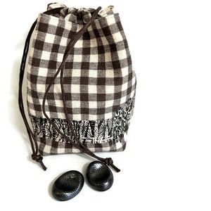 Pode incluir: Uma bolsa de cordão de gingham marrom e branco com uma borda floral preta e branca. A bolsa é amarrada com um cordão marrom e tem dois botões de cerâmica preta na superfície branca abaixo.