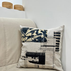 May include: A beige linen throw pillow with a black and white geometric design. The design features a large square with a smaller square inside, and a smaller square with a floral pattern. The pillow is stitched with black thread.