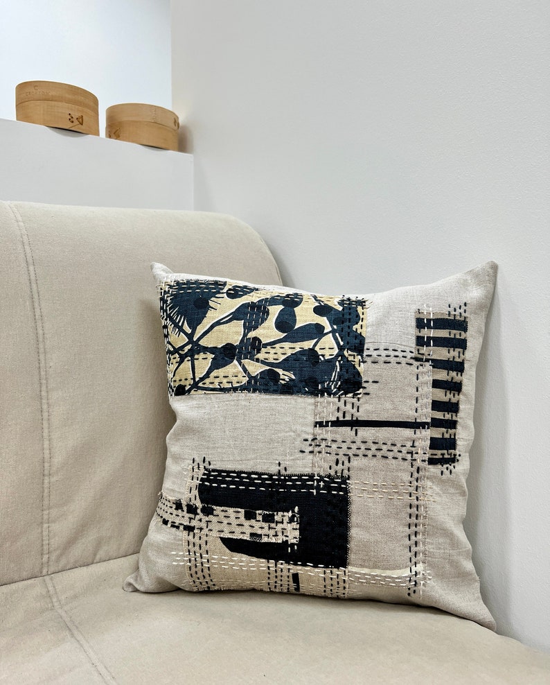 May include: A beige linen throw pillow with a black and white geometric design. The design features a large square with a smaller square inside, and a smaller square with a floral pattern. The pillow is stitched with black thread.