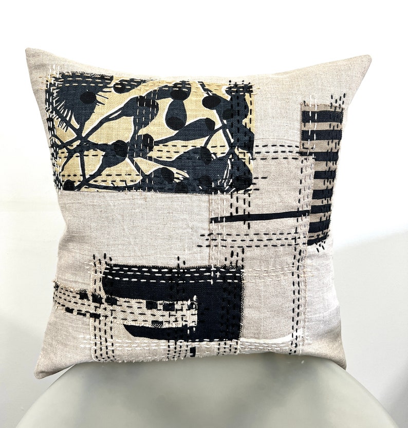 May include: A beige linen pillow with black and white geometric patterns and stitching. The pillow has a patchwork design with different fabrics and textures.