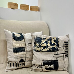 May include: Two decorative throw pillows with black and white geometric designs on a beige linen fabric. The pillows are sitting on a beige couch.