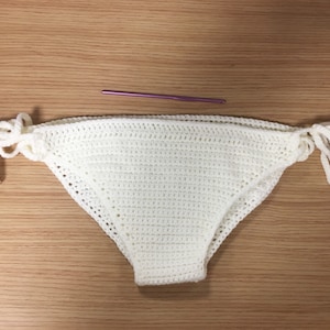 May include: A handmade, white crochet bikini bottom with tie sides. The crochet work is detailed, and the ties are long and delicate. A purple crochet hook rests above the bikini bottom on a wooden surface.