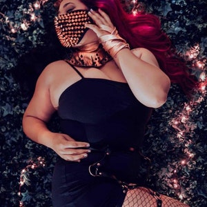 May include: A black dress with spaghetti straps, paired with a studded face mask and choker. The ensemble includes a leather belt with metal hardware and fishnet stockings. The model's hair is a vibrant red, and the accessories have a metallic gold finish.