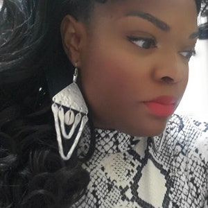 Puede incluir: Una mujer con un top de estampado de piel de serpiente blanco y negro con pendientes de plata colgantes. Los pendientes tienen forma triangular con un acabado de metal martillado y un acento redondo blanco.