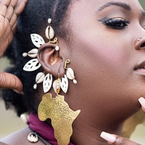 May include: A woman wearing a gold earring shaped like the continent of Africa. The earring is attached to a smaller earring with white shell-like pieces.