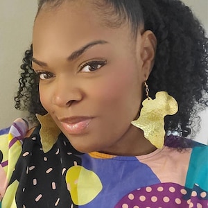 May include: A woman with curly hair is wearing gold earrings shaped like the continent of Africa. She is wearing a colourful shirt with a geometric pattern.