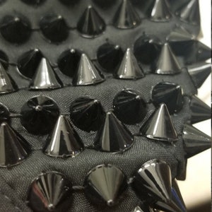 May include: Close-up of a black fabric with rows of shiny, cone-shaped and rounded studs. The studs are evenly spaced and create a textured, edgy design. The fabric appears to be a dark gray color.