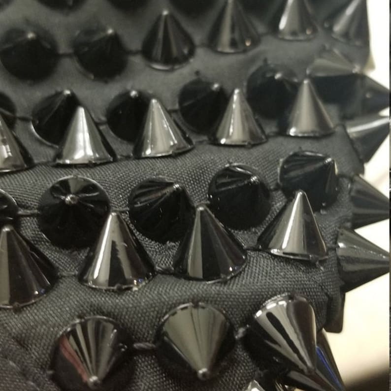May include: Close-up of a black fabric with rows of shiny, cone-shaped and rounded studs. The studs are evenly spaced and create a textured, edgy design. The fabric appears to be a dark gray color.