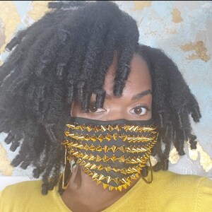 May include: Black face mask adorned with gold-colored spikes. The mask covers the lower face and is set against a background of black hair and a yellow top. The spikes are evenly spaced and create a bold, edgy design.