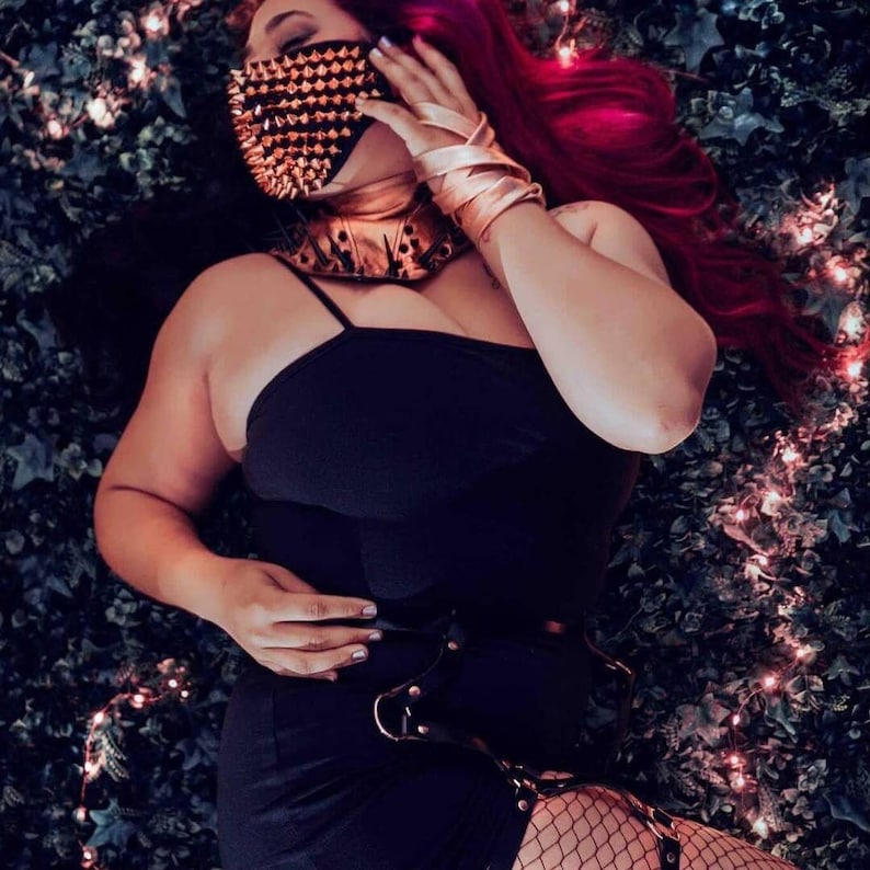 May include: A black dress with spaghetti straps, paired with a studded face mask and choker. The ensemble includes a leather belt with metal hardware and fishnet stockings. The model's hair is a vibrant red, and the accessories have a metallic gold finish.