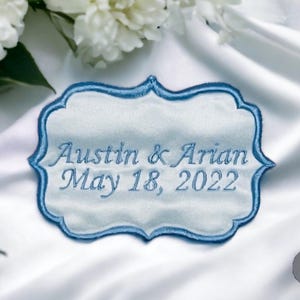 May include: A light blue embroidered wedding favor with the names "Austin & Arian" and the date "May 18, 2022". The embroidered label has a decorative border and is set against a white background.