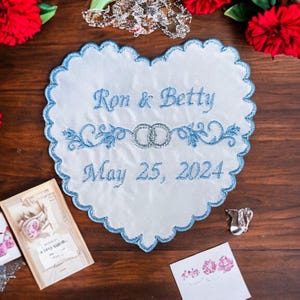 May include: A white heart-shaped embroidered wedding keepsake with blue scalloped edges. The text "Ron & Betty" and "May 25, 2024" are embroidered in blue. Two silver rings are embroidered in the center. Red flowers and other wedding-related items surround the keepsake.