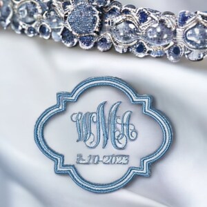 May include: A light blue embroidered emblem with the initials "WMH" and the date "2-10-2023". The emblem has a decorative border and is set against a white satin background. A jeweled tiara is visible in the background.
