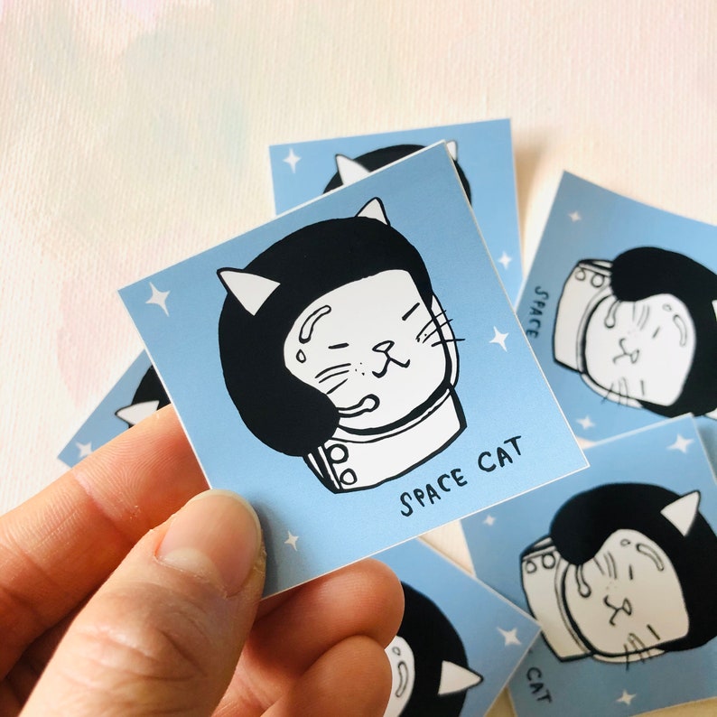 K&ouml;nnte beinhalten: Ein blauer Aufkleber mit einer wei&szlig;en Katze, die einen schwarzen Astronautenhelm tr&auml;gt. Die Katze hat einen &uuml;berraschten Ausdruck und der Text "SPACE CAT" ist unter der Katze gedruckt.