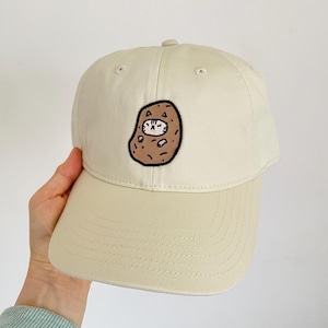 May include: A beige baseball cap with a brown and white embroidered cat inside a potato design.