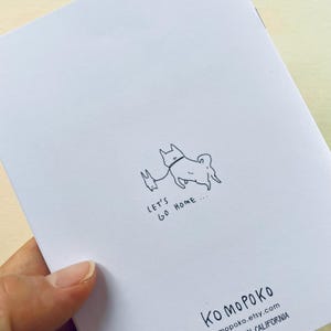 May include: A white card with a black and white illustration of two dogs walking on a leash. The text "Let's go home..." is written below the illustration. The text "KO MOPOKO" is written in the bottom right corner of the card.