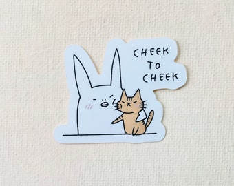 Puede incluir: Una pegatina blanca con una ilustración de dibujos animados de un gran conejo blanco y un pequeño gato atigrado naranja. El gato está tocando la mejilla del conejo. El texto "CHEEK TO CHEEK" está escrito en negro.