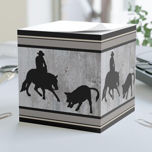 Può includere: Un blocco note quadrato con un design grigio e nero che presenta la silhouette di un cowboy a cavallo che guida un vitello. Il blocco note ha un foglio superiore bianco.