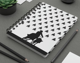 May include: A white spiral bound notebook with a black and white silhouette pattern of a person riding a horse. The notebook has a black border.