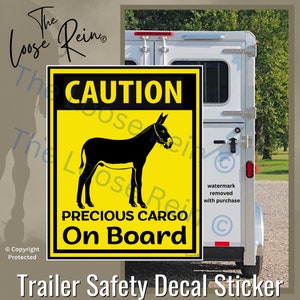 May include: Yellow caution sign with black text that reads "CAUTION PRECIOUS CARGO On Board". A black silhouette of a donkey is in the center of the sign. The sign is attached to the back of a white horse trailer.
