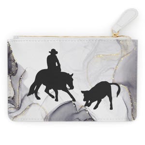 May include: A white zippered pouch with a marbleized design in shades of gray and gold. The pouch features a black silhouette of a cowboy riding a horse and a calf.