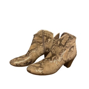 May include: A pair of ankle boots with a snakeskin pattern in shades of tan and brown. The boots have a side zip and a small heel. The overall style is western-inspired.